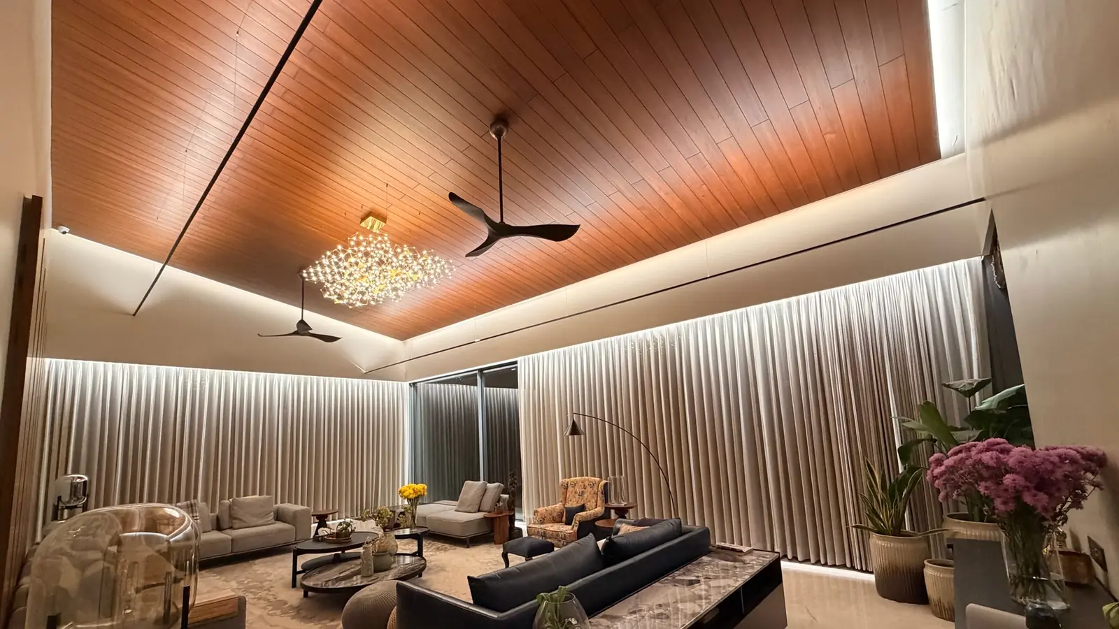 A living room in the evening: a timber ceiling with a lit cove, long curtains washed with light, and a chandelier over the seating.